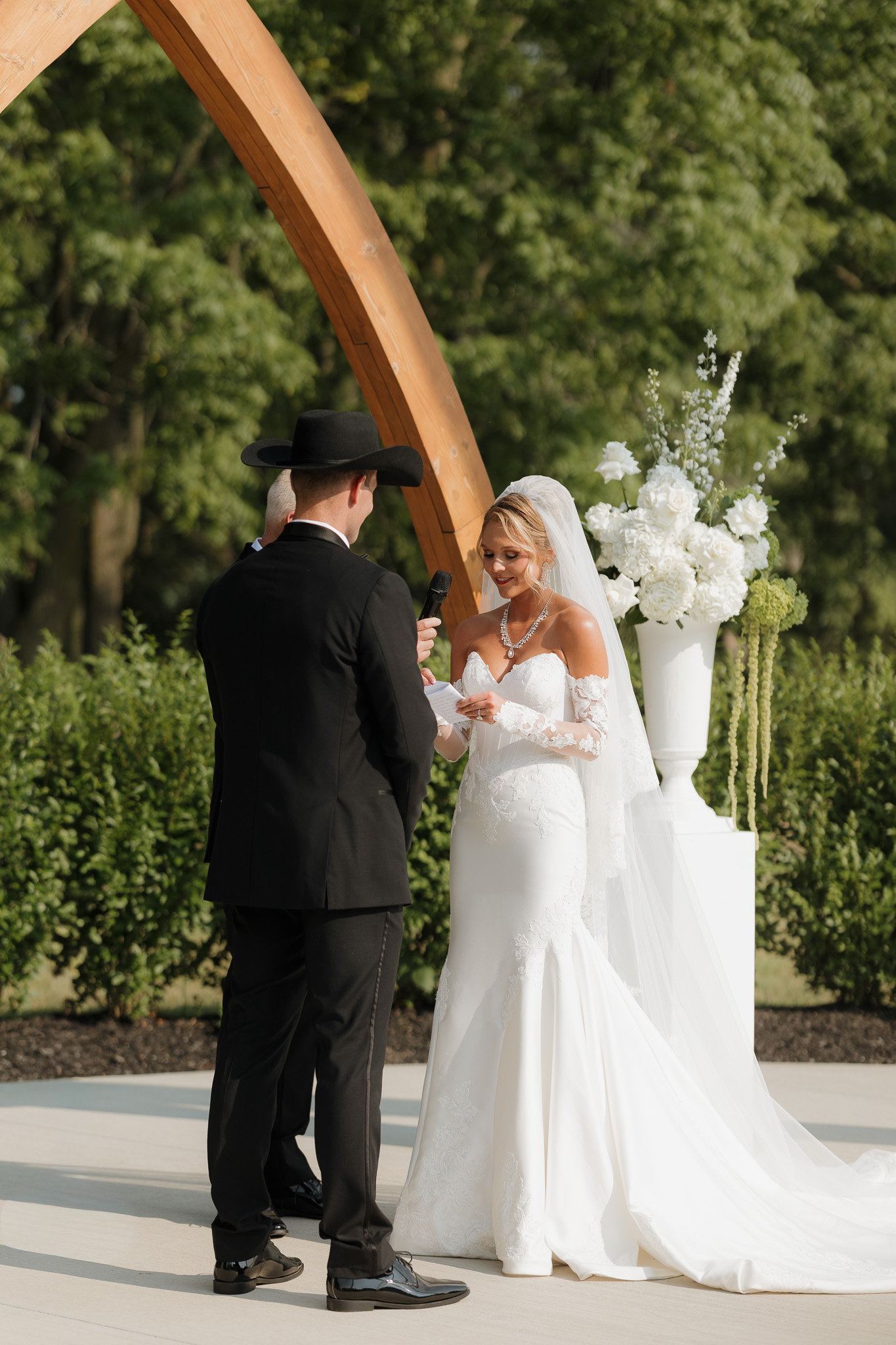 Bride and groom exchanging vows beneath the Grand Arches surrounded by white floral arrangements during their outdoor ceremony at The Wells Estate.