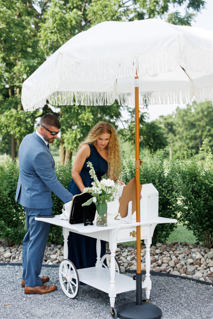 Guests visiting a white dessert cart beneath a fringed umbrella during an outdoor summer wedding reception at The Wells Estate.