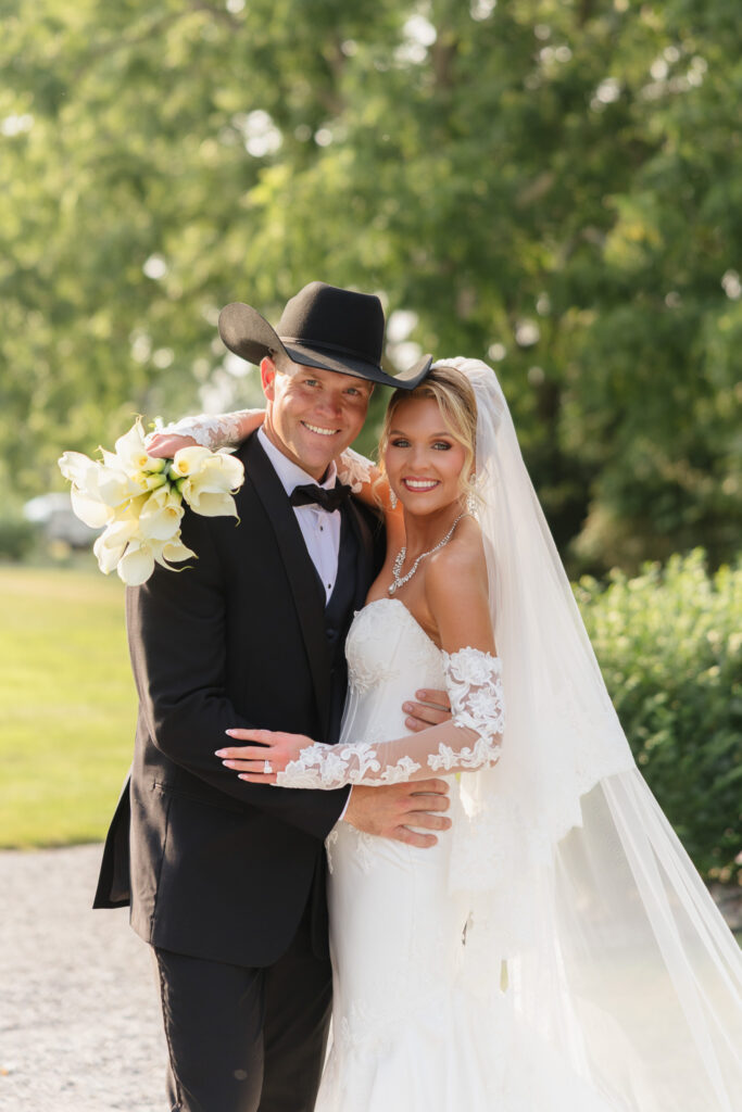Bride in a lace wedding gown and groom in a black tuxedo and cowboy hat posing together outdoors after their summer wedding ceremony at The Wells Estate.