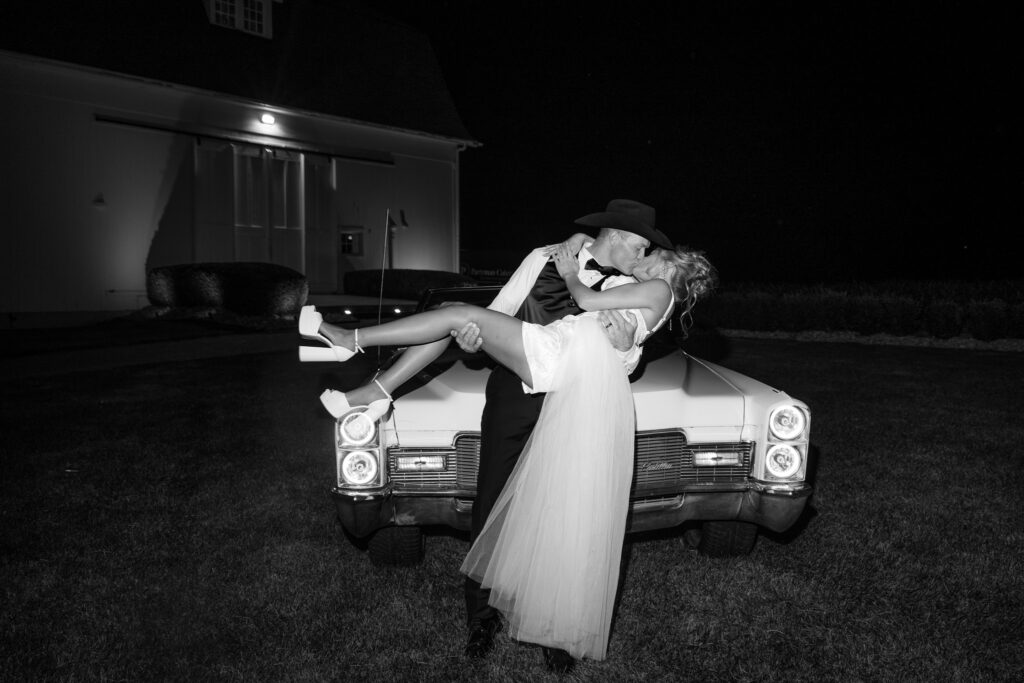 Black-and-white nighttime wedding portrait of the groom dipping and kissing the bride in front of a vintage white convertible at The Wells Estate.