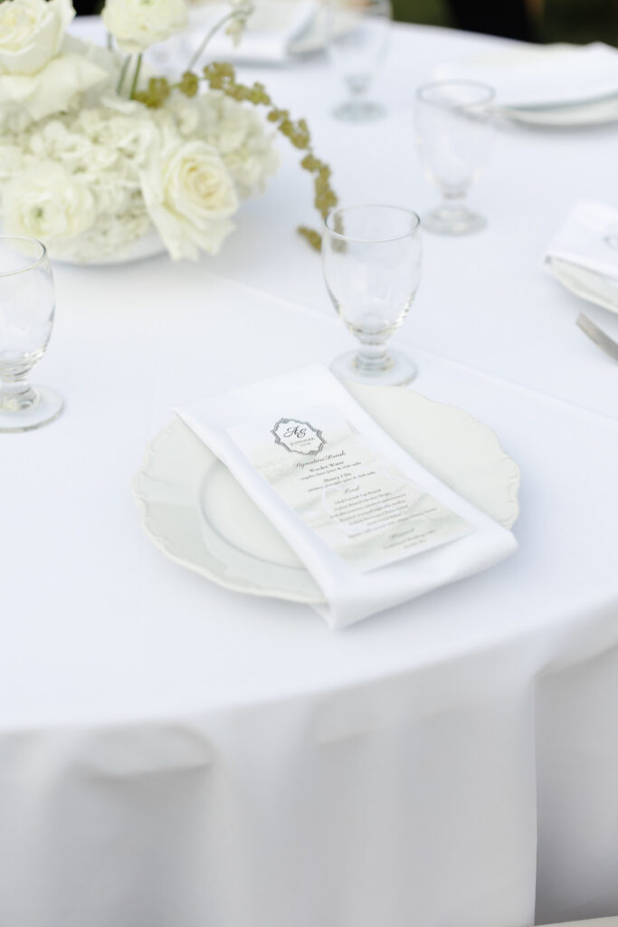 Elegant white wedding place setting with a personalized menu, white linen napkin, glassware, and white floral centerpiece at The Wells Estate.