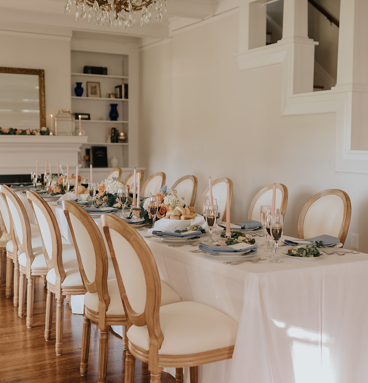 A long formal dinner table at The Wells Estate, styled with neutral linens, soft peach florals, and gold-rimmed chairs under a chandelier for an intimate and refined celebration.